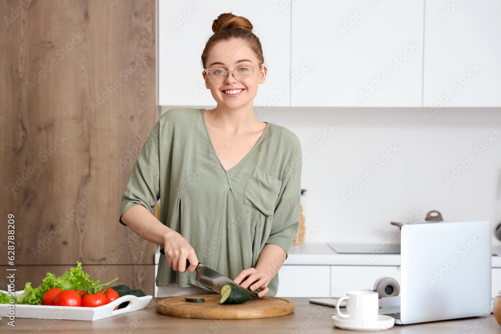 Beautiful young woman cutting cucumber at table with modern laptop in light kitchen Beautiful young woman cutting cucumber at table with modern laptop in light kitchen