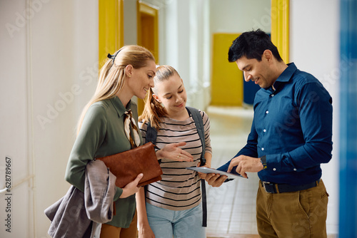 Male principal using touchpad with high school student and her mother in hallway.