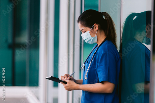 Asian nurse wearing surgical mask and stethoscope holding digital tablet in hospital