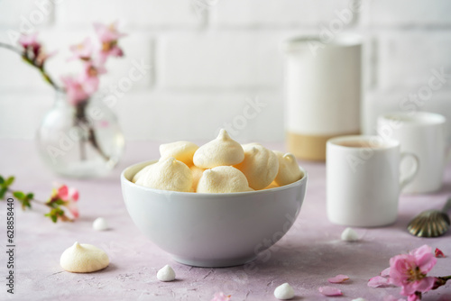 Small meringues in a white bowl over light pink background decorated with spring peach tree flowers, two cup of coffee behind