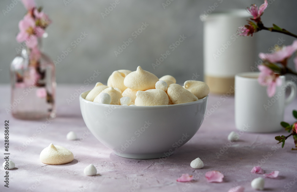 Small meringues in a white bowl over light pink and grey concrete background decorated with spring peach flowers, cup of coffee, creamer behind - obrazy, fototapety, plakaty 