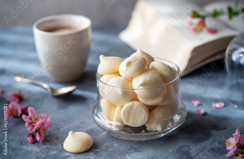 Small meringues in a glass bowl over grey concrete background decorated with spring peach flowers, cups of coffee, book behind