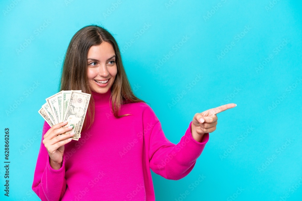 Young caucasian woman taking a lot of money isolated on blue background pointing finger to the side and presenting a product Young caucasian woman taking a lot of money isolated on blue background pointing finger to the side and presenting a product