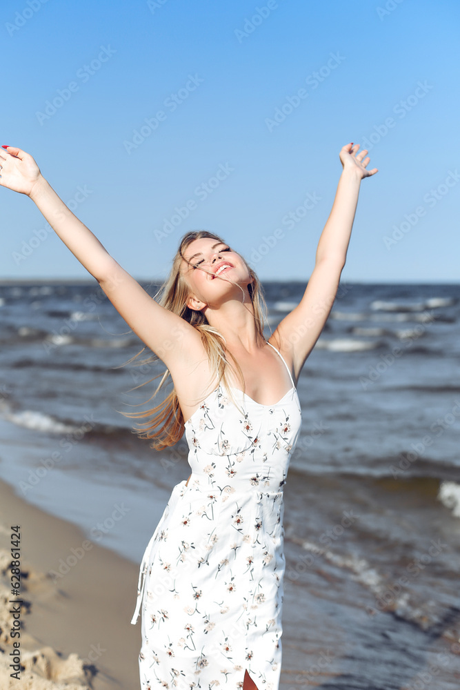 Happy blonde beautiful woman on the ocean beach standing in a white summer dress, raising hands Happy blonde beautiful woman on the ocean beach standing in a white summer dress, raising hands