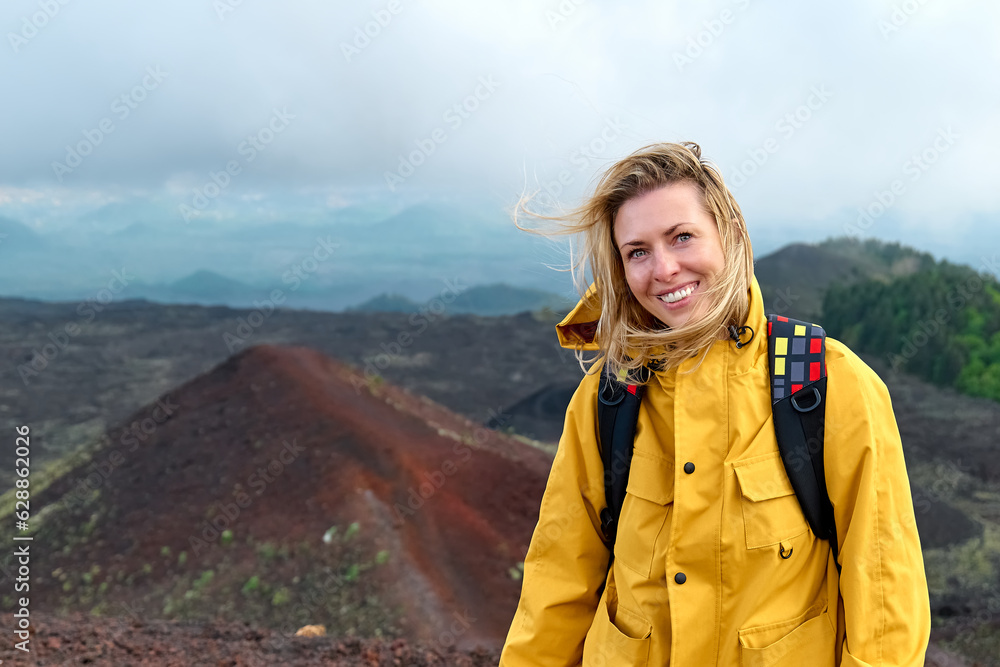 Hiking on tallest volcano in Continental Europe - Etna. Young smiling woman in yellow raincoat in the mountain peak of panoramic view of Mount Etna in a windy day.