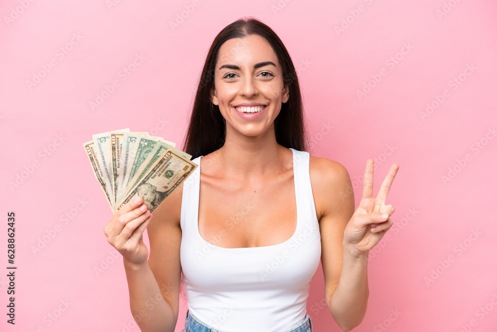 Young caucasian woman taking a lot of money isolated on pink background smiling and showing victory sign Young caucasian woman taking a lot of money isolated on pink background smiling and showing victory sign