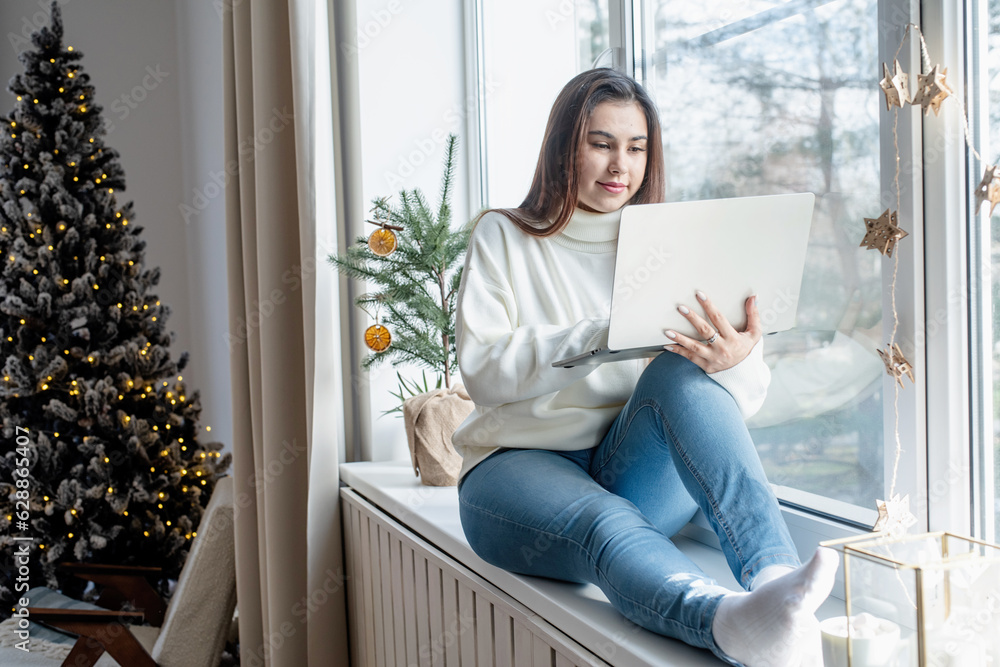Woman in warm white winter sweater sitting at windowsill at home at christmas eve working on laptop Woman in warm white winter sweater sitting at windowsill at home at christmas eve working on laptop