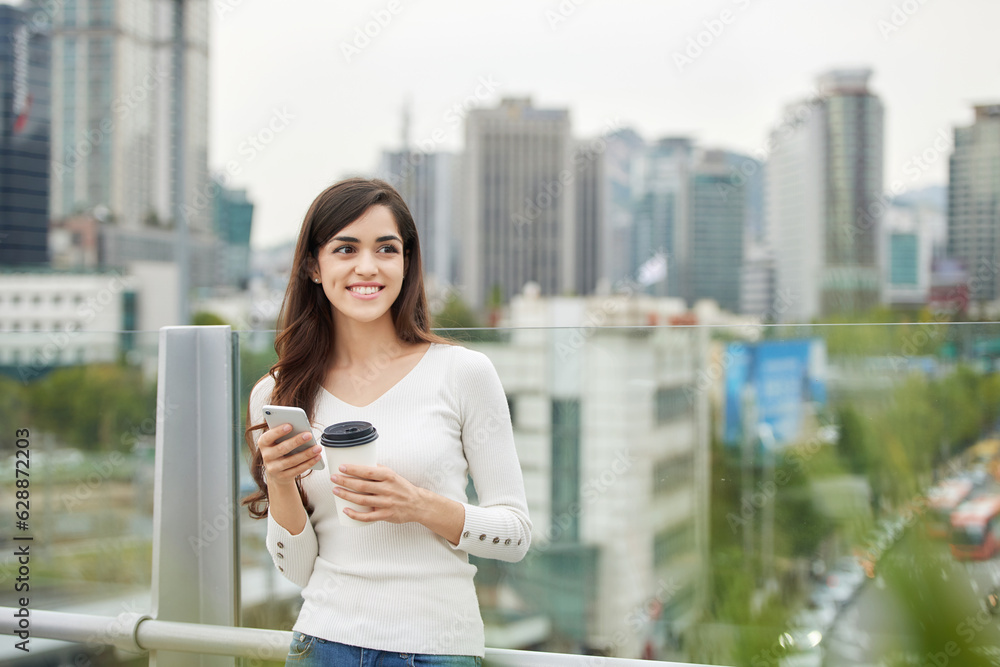 A young woman is resting on a land bridge with coffee and a smartphone in the background of a busy street during the day.