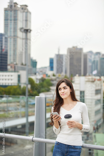 A young woman is resting on a land bridge with coffee and a smartphone in the background of a busy street during the day.