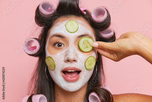 Joyful girl with wavers on her black hair making facial procedures wth nutrition mask and fresh cucumbers, face treatment concept, copy space