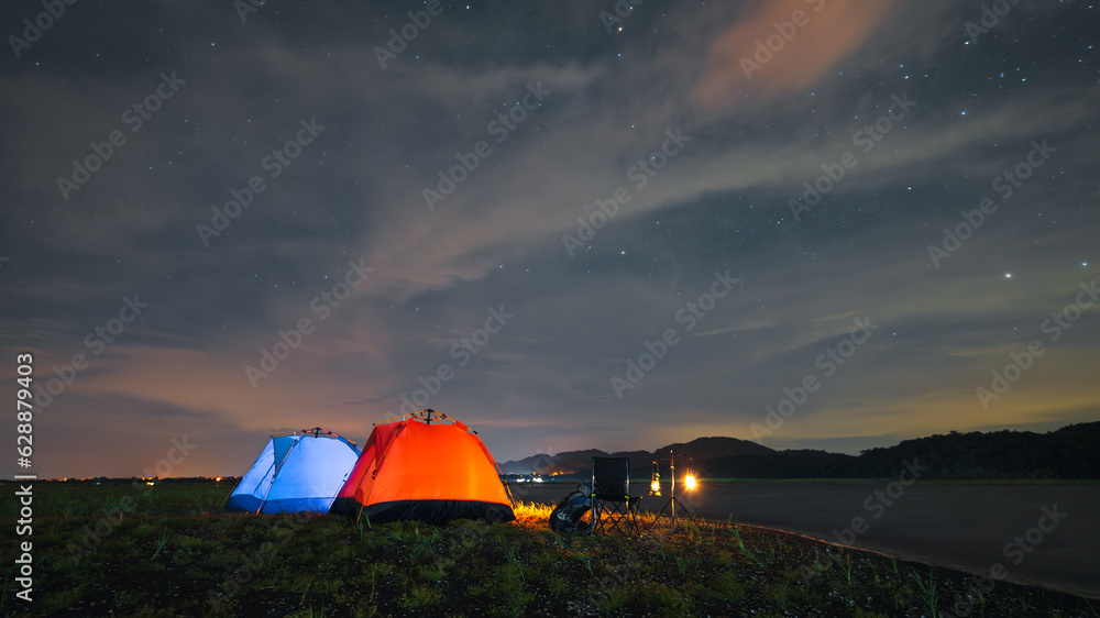 riverside camping, blue and orange riverside tent camping with luggage, bonfire, evening camping activities The atmosphere of camping at night when the sky is full of stars.