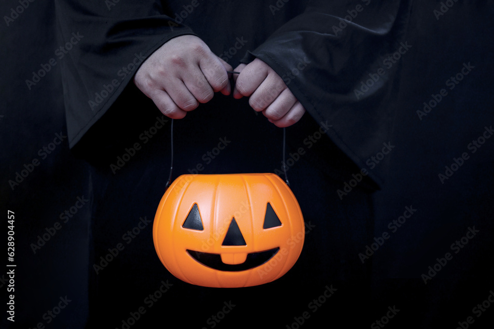 Halloween pumpkin in hand of man in spooky black costume standing in dark background, sorcerer wizard holding orange pumpkin going to celebrate Halloween party, playing trick or treat. Halloween pumpkin in hand of man in spooky black costume standing in dark background, sorcerer wizard holding orange pumpkin going to celebrate Halloween party, playing trick or treat.
