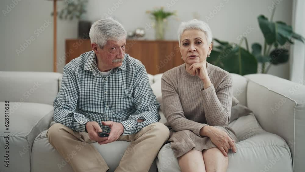 Senior couple wife and husband watches TV sits on sofa at home. Man switches channels uses remote control talks with woman discusses their choose. Family entertainment, relax, spending time together. Senior couple wife and husband watches TV sits on sofa at home. Man switches channels uses remote control talks with woman discusses their choose. Family entertainment, relax, spending time together.