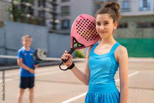 Cheerful young girl standing with padel racket and with blurred friend on sports ground