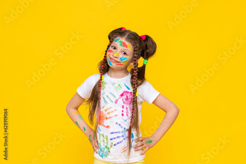 Happy little girl painted with colorful paints. A child with a stained face in paints, enjoys drawing. Art art for children and schoolchildren. Yellow isolated background.