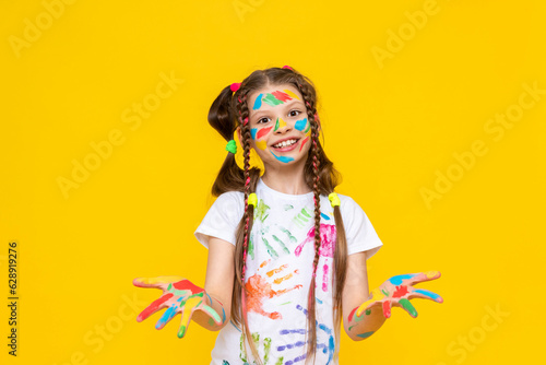 A little girl stained with multicolored paints. A happy, beautiful young girl painted with artistic paints. The child shows his smeared palms in paint and smiles. Yellow isolated background.