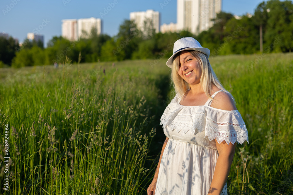 Copy space. Cute blond woman squinting stands in the summer in a green field in the rays of the setting sun. A squinty woman looks at the camera with one eye covered against a background of greenery. Copy space. Cute blond woman squinting stands in the summer in a green field in the rays of the setting sun. A squinty woman looks at the camera with one eye covered against a background of greenery.