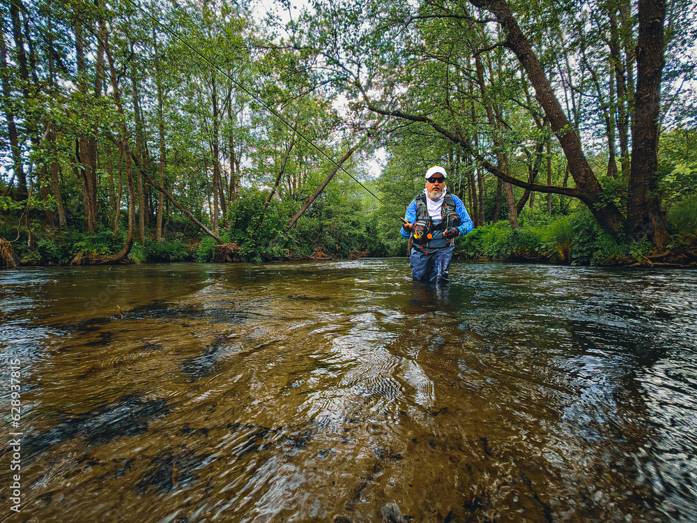 Man with fishing rod in green forest, standing by river, looking at camera Man with fishing rod in green forest, standing by river, looking at camera
