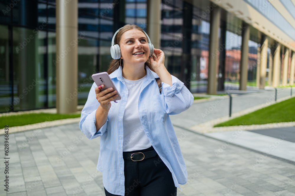 Young happy woman wearing suit using wireless earphones holding smartphone talking on mobile phone having chat on cellphone walking on urban city street. High quality photo Young happy woman wearing suit using wireless earphones holding smartphone talking on mobile phone having chat on cellphone walking on urban city street. High quality photo