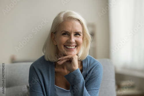 Canvas Print Cheerful pretty blonde senior woman looking at camera, smiling with healthy white teeth, laughing, posing for shooting on sofa, touching chin