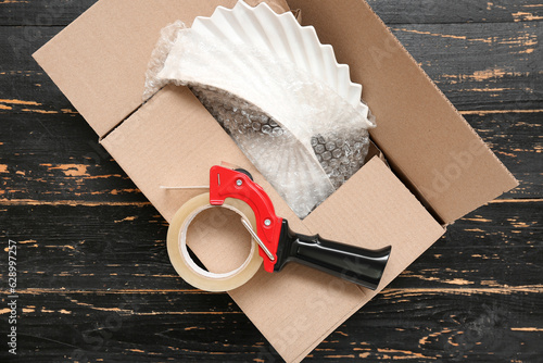 Packing tape dispenser and cardboard box on black wooden background