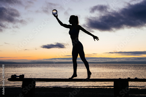 Silhouette of young woman doing workout on a bench with crystal ball(s), in front of ocean at sunrise or sunset