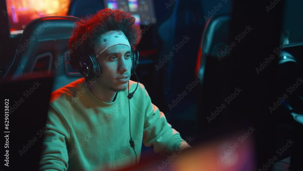 Medium shot of young multiethnic guy with curly hair, headband and headset sitting in cyberclub at night, playing shooter video game and sipping energy drink from can Medium shot of young multiethnic guy with curly hair, headband and headset sitting in cyberclub at night, playing shooter video game and sipping energy drink from can
