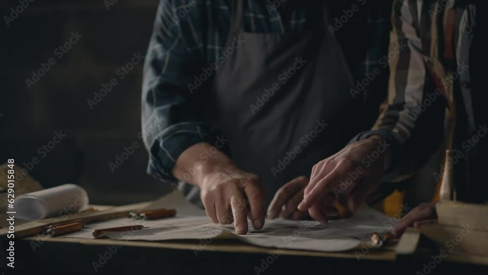 Handicraft Two colleagues workers studying blueprints of wooden parts for manufacturing At the workplace A young professional performs the task of making parts In a production room, small business Handicraft Two colleagues workers studying blueprints of wooden parts for manufacturing At the workplace A young professional performs the task of making parts In a production room, small business