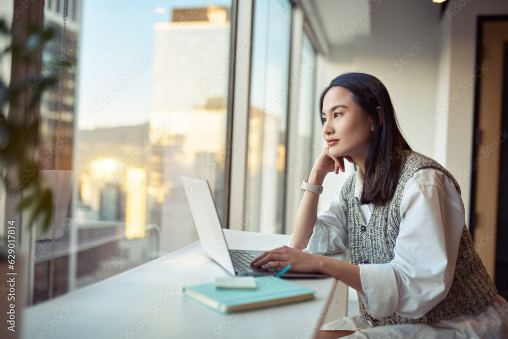 Young pretty pensive Asian professional business woman employee or student working on laptop in corporate office sitting at desk looking at window thinking of new ideas or dreaming, contemplating. Young pretty pensive Asian professional business woman employee or student working on laptop in corporate office sitting at desk looking at window thinking of new ideas or dreaming, contemplating.