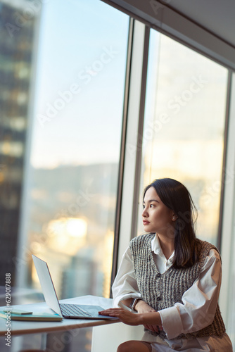 Young pretty pensive Asian professional business woman employee or student working on laptop in corporate office sitting at desk looking at window thinking of new ideas or dreaming, vertical shot. Free Download Young pretty pensive Asian professional business woman employee or student working on laptop in corporate office sitting at desk looking at window thinking of new ideas or dreaming, vertical shot. - GFXLoad