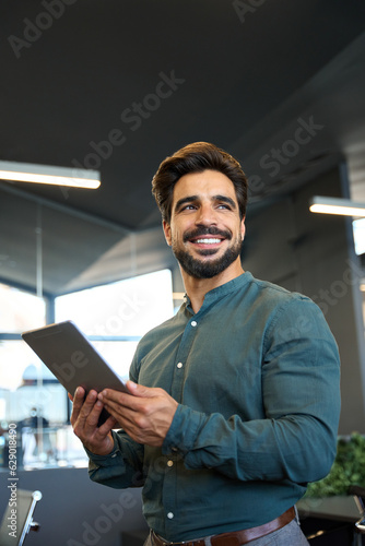 Smiling young Latin business man entrepreneur using tablet standing in office at work. Happy male professional executive manager holding tab computer looking away and thinking of tech data. Vertical