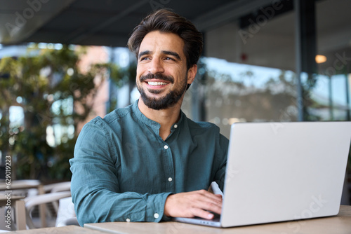 Fototapeta Smiling young Latin business man using laptop sitting outdoor