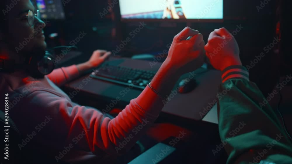 Close-up rear slowmo shot of two young male friends playing first person shooter game together in cyber cafe, winning and bumping fists Close-up rear slowmo shot of two young male friends playing first person shooter game together in cyber cafe, winning and bumping fists