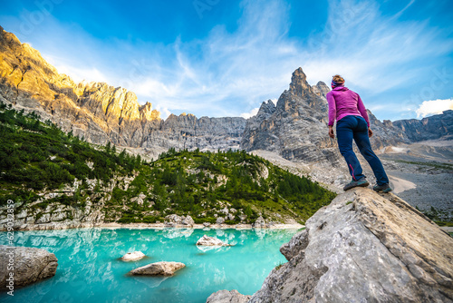 Athletic woman stands on big boulder at turquoise Sorapis lake with dito di dio in the background in the evening. Lake Sorapis, Dolomites, Belluno, Italy, Europe.