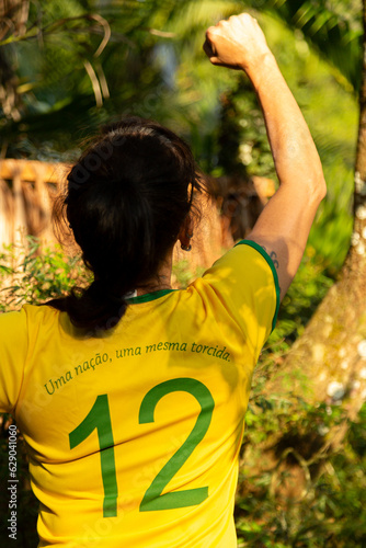 Torcedora brasileira de costas com a blusa verde amarela numero 12, comemorando o gol com soco no ar e ao fundo a natureza.