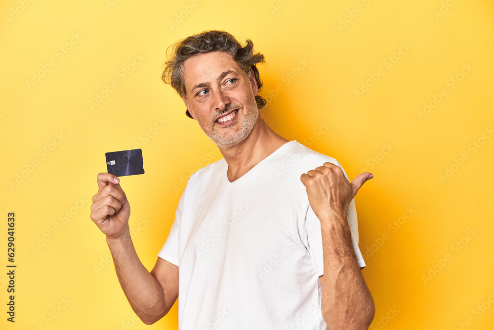 Man holding a credit card, yellow studio backdrop points with thumb finger away, laughing and carefree. Man holding a credit card, yellow studio backdrop points with thumb finger away, laughing and carefree.