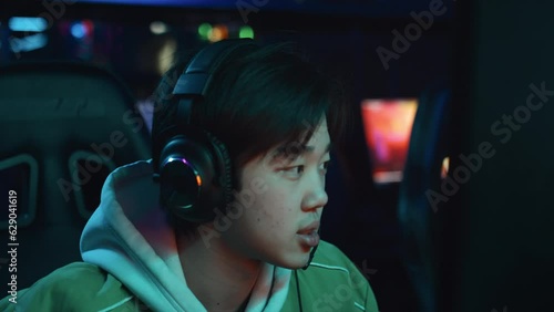 Close-up shot of face of young Chinese male gamer in hooded sweatshirt and headset playing multiplayer shooter video game in cyberclub and talking into microphone to coordinate with team photo