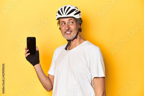 Cyclist man showing phone on yellow backdrop looks aside smiling, cheerful and pleasant.