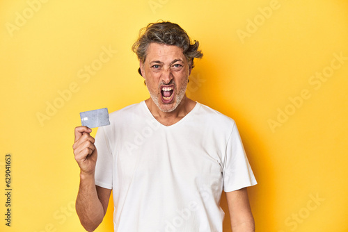 Man holding a credit card, yellow studio backdrop screaming very angry and aggressive.