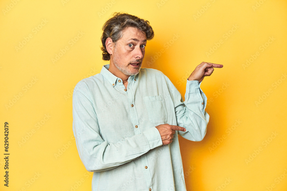 Middle-aged man posing on a yellow backdrop shocked pointing with index fingers to a copy space. Middle-aged man posing on a yellow backdrop shocked pointing with index fingers to a copy space.