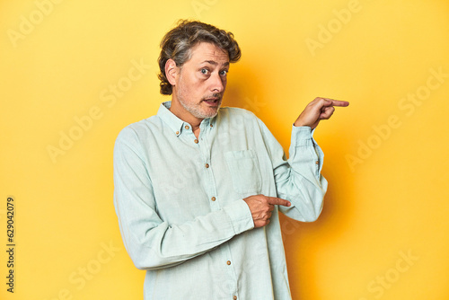 Middle-aged man posing on a yellow backdrop shocked pointing with index fingers to a copy space.
