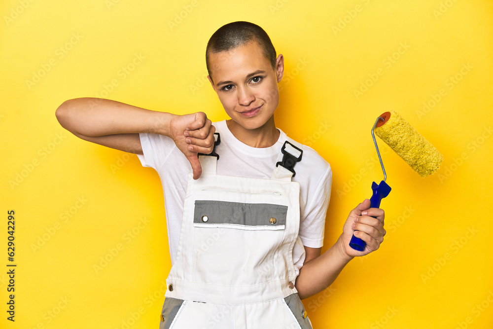 Painter woman, shaved head, with yellow roller in studio showing a dislike gesture, thumbs down. Disagreement concept. Painter woman, shaved head, with yellow roller in studio showing a dislike gesture, thumbs down. Disagreement concept.