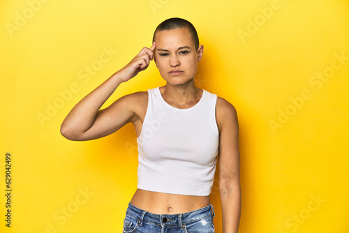 Shaved head woman in white tank top, yellow backdrop pointing temple with finger, thinking, focused on a task.