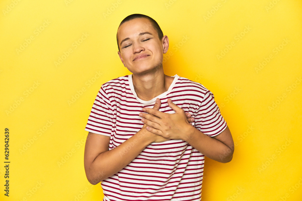 Shaved head woman in red striped tee, yellow backdrop laughing keeping hands on heart, concept of happiness. Shaved head woman in red striped tee, yellow backdrop laughing keeping hands on heart, concept of happiness.