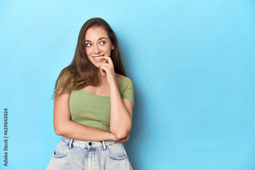 Young Caucasian woman in a green top on a blue backdrop relaxed thinking about something looking at a copy space. Young Caucasian woman in a green top on a blue backdrop relaxed thinking about something looking at a copy space.