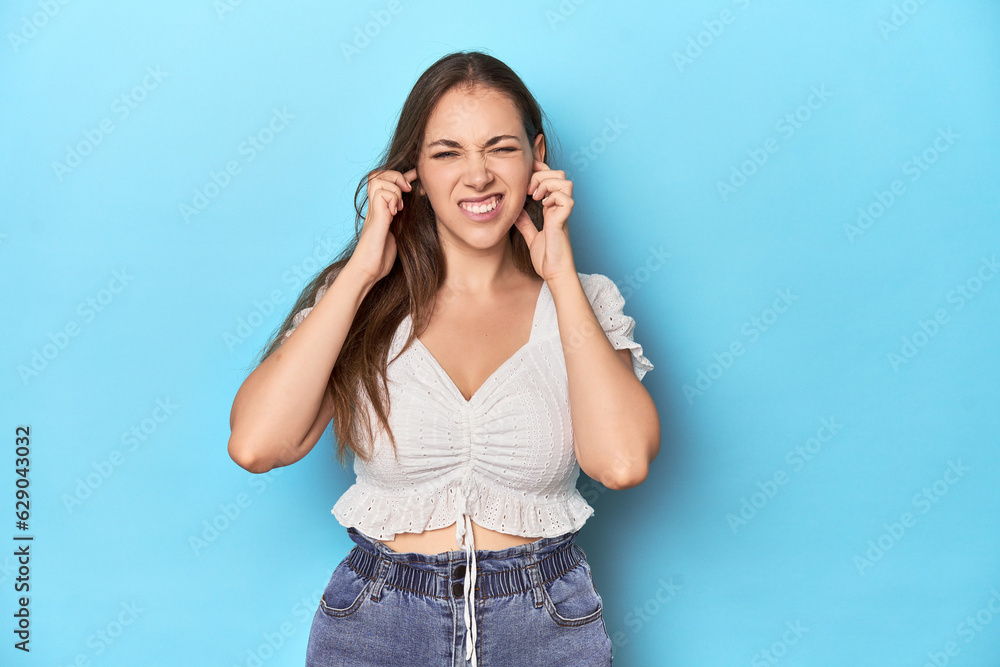 Stylish young woman in white blouse on a blue studio backdrop covering ears with hands. Stylish young woman in white blouse on a blue studio backdrop covering ears with hands.