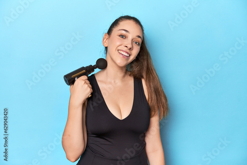 Fitness woman holding electric massager in a blue studio setting.