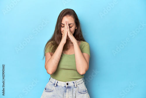 Young Caucasian woman in a green top on a blue backdrop holding hands in pray near mouth, feels confident.