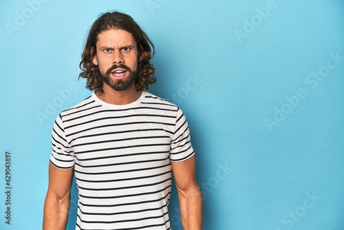 Bearded man in a striped shirt, blue backdrop shouting very angry, rage concept, frustrated.