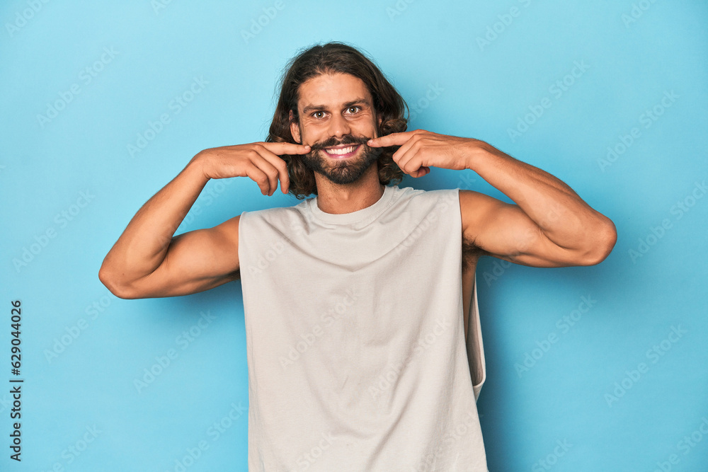 Bearded man in a tank top, blue backdrop smiles, pointing fingers at mouth. Bearded man in a tank top, blue backdrop smiles, pointing fingers at mouth.